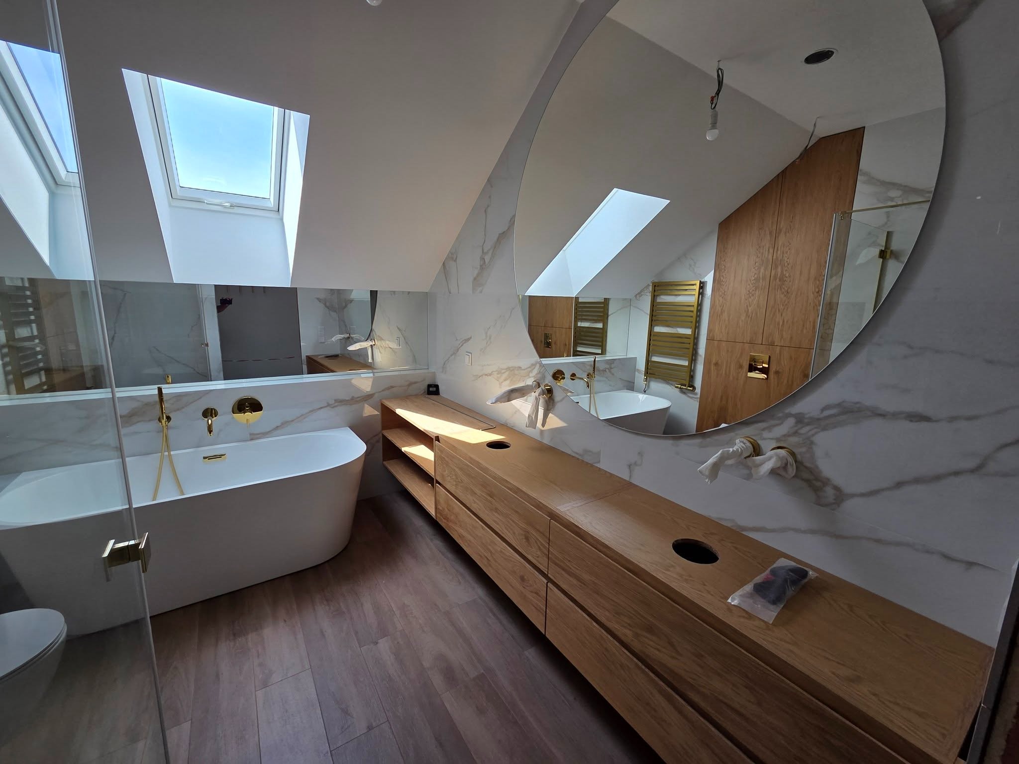 Jasna łazienka na poddaszu z oknem dachowym, marmurowymi płytkami i drewnianą zabudową umywalki. Duże, okrągłe lustro odbija wnętrze. Widać wannę i złote akcenty.
