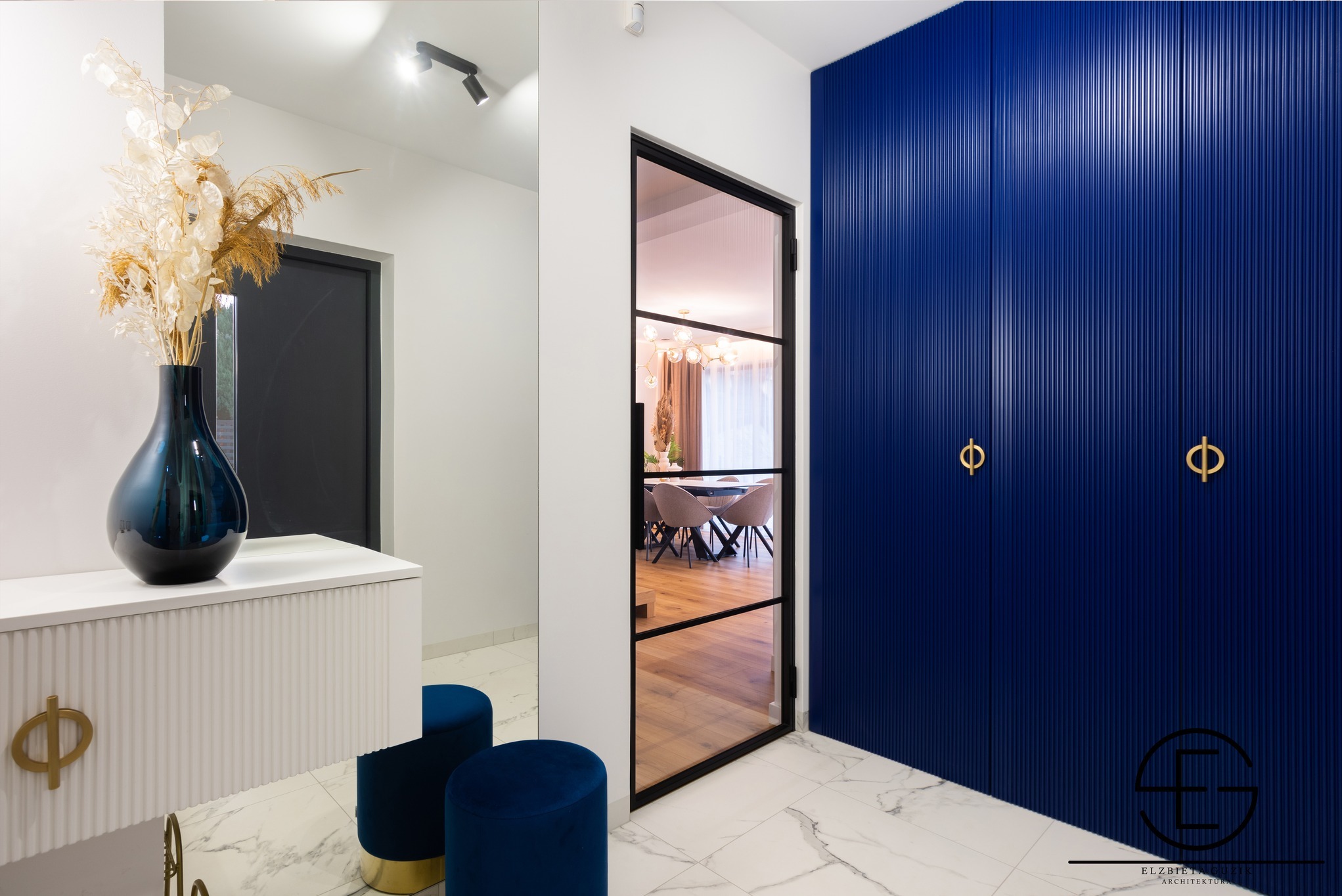 Elegancki przedpokój: biała komoda z dekoracyjnym wazonem, lustro, granatowe pufy i szafa w odcieniach granatu z ryflowanymi frontami oraz złote uchwyty.