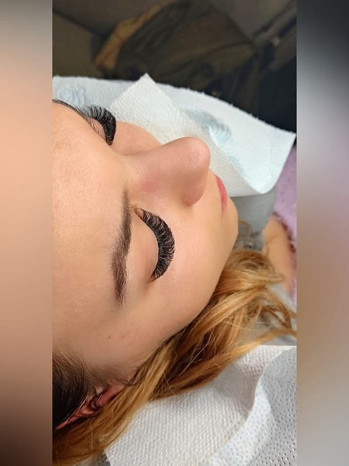 Klientka z przedłużonymi rzęsami leży na podkładzie, widok z bliska na oko z gęstymi, czarnymi rzęsami. Blond włosy, delikatny makijaż ust.