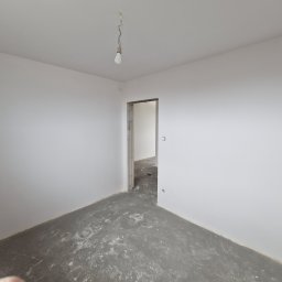 S&M Solutions Szymon Maszota - Pusty pokój w trakcie remontu: białe ściany, surowy beton na podłodze, wisząca żarówka. Widoczne ślady prac wykończeniowych i przygotowania do dalszych etapów.