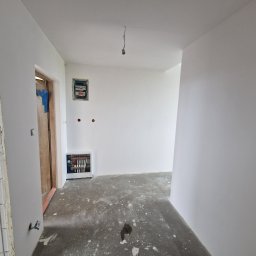 S&M Solutions Szymon Maszota - Wnętrze po szpachlowaniu i malowaniu na biało. Widoczna instalacja grzewcza, rozdzielnia elektryczna i surowa ościeżnica drzwiowa.