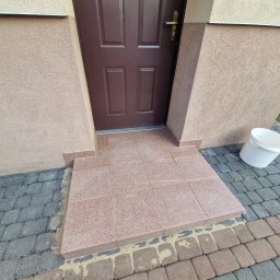 S&M Solutions Szymon Maszota - Wykończone płytkami schody wejściowe do domu, z widocznym brązowym wejściem i kostką brukową. Białe wiadro stoi obok schodów.