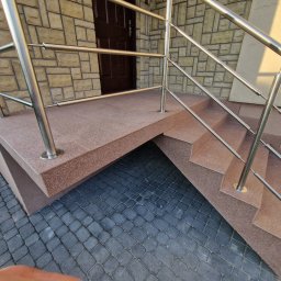 S&M Solutions Szymon Maszota - Zewnętrzne schody z terakoty z metalową balustradą, prowadzące do drzwi wejściowych. Kamienna elewacja budynku w tle i kostka brukowa na dole.