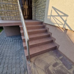 S&M Solutions Szymon Maszota - Zewnętrzne schody wejściowe wyłożone brązowymi płytkami, z metalową barierką, prowadzące do ciemnych drzwi wejściowych. Widoczna elewacja z kamienia i faktura tynku.