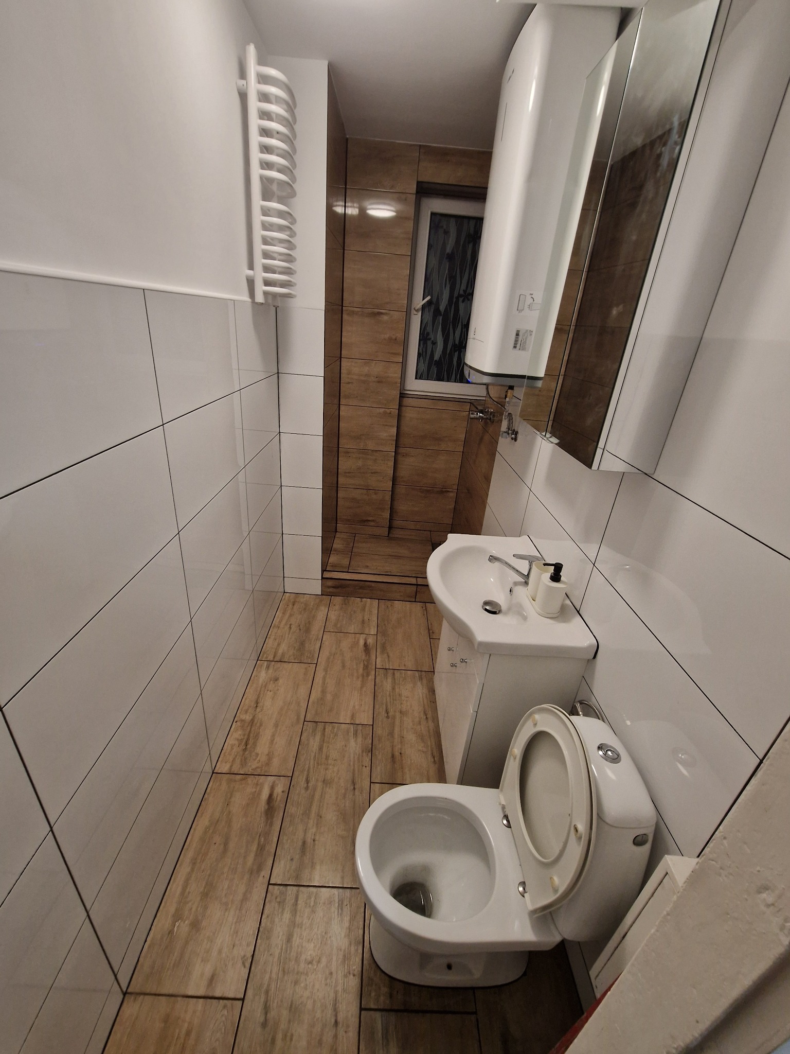 Wnętrze łazienki po modernizacji: białe płytki ścienne, drewnopodobne płytki podłogowe, umywalka z szafką, toaleta i grzejnik drabinkowy. Widok z góry, wąskie ujęcie.