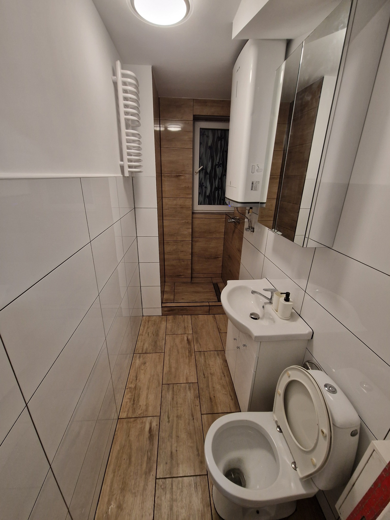 Wąska łazienka z białymi płytkami i drewnopodobną podłogą. Umywalka z szafką, toaleta i grzejnik drabinkowy. W tle kabina prysznicowa z drewnianymi panelami.