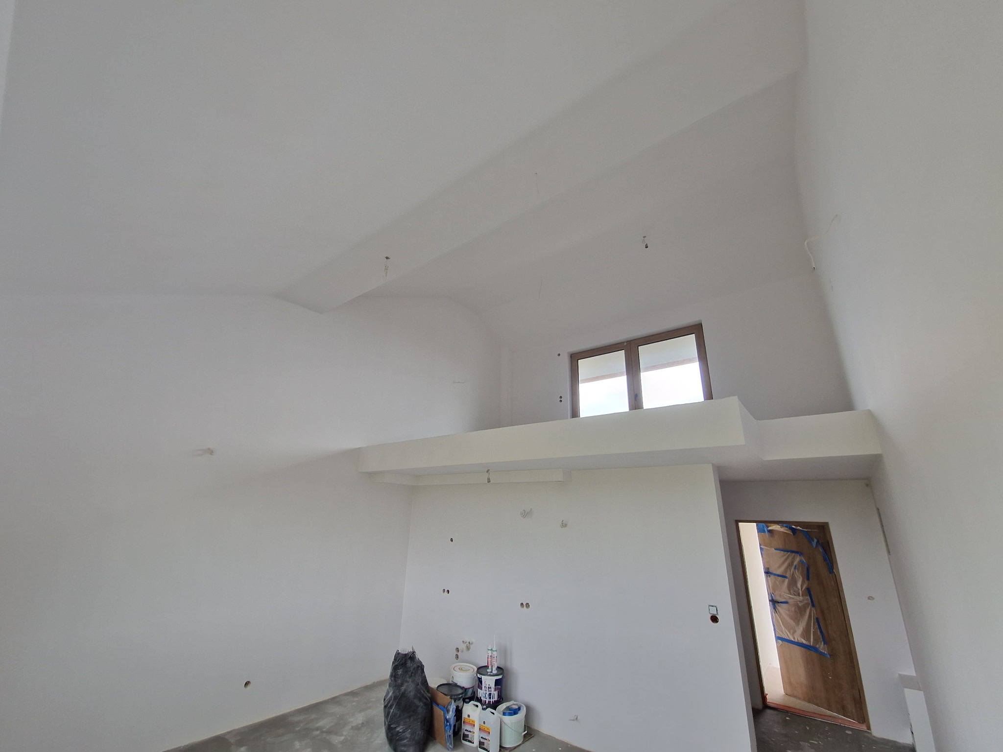 Wnętrze w bieli: świeżo pomalowane ściany z widocznymi punktami po instalacjach, okno w górnej części, drzwi zabezpieczone folią, farby i narzędzia malarskie.