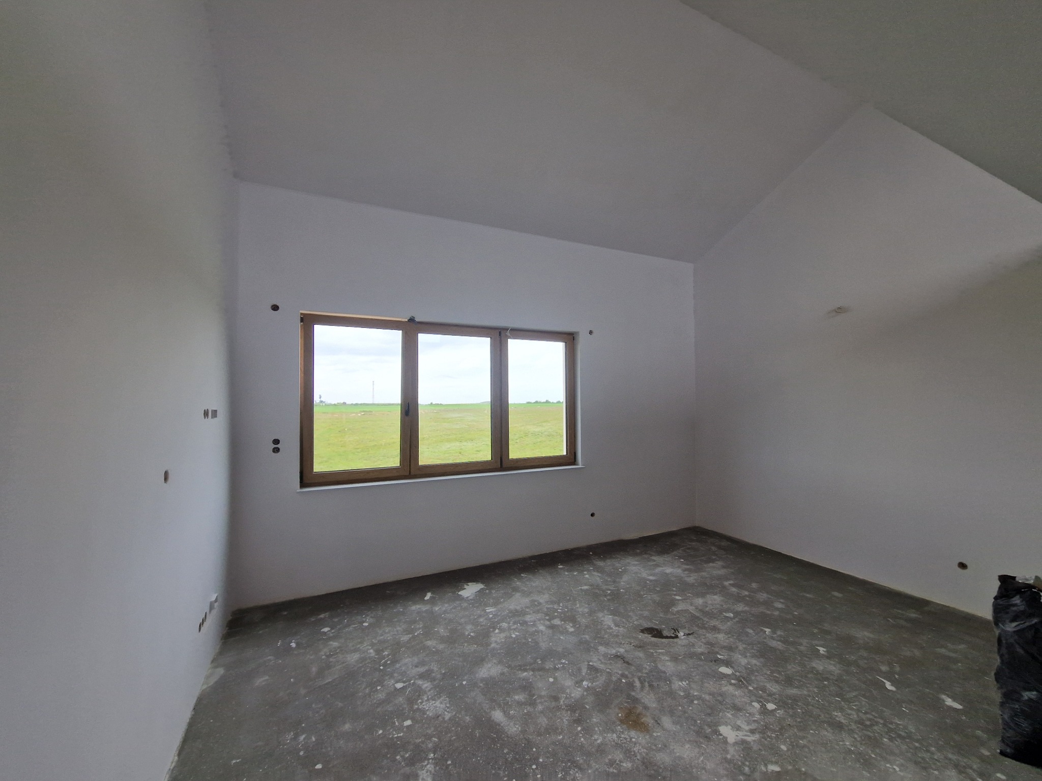 Surowe wnętrze z oknem na krajobraz. Szpachlowane ściany, przygotowane do dalszych prac wykończeniowych. Widoczne punkty elektryczne.