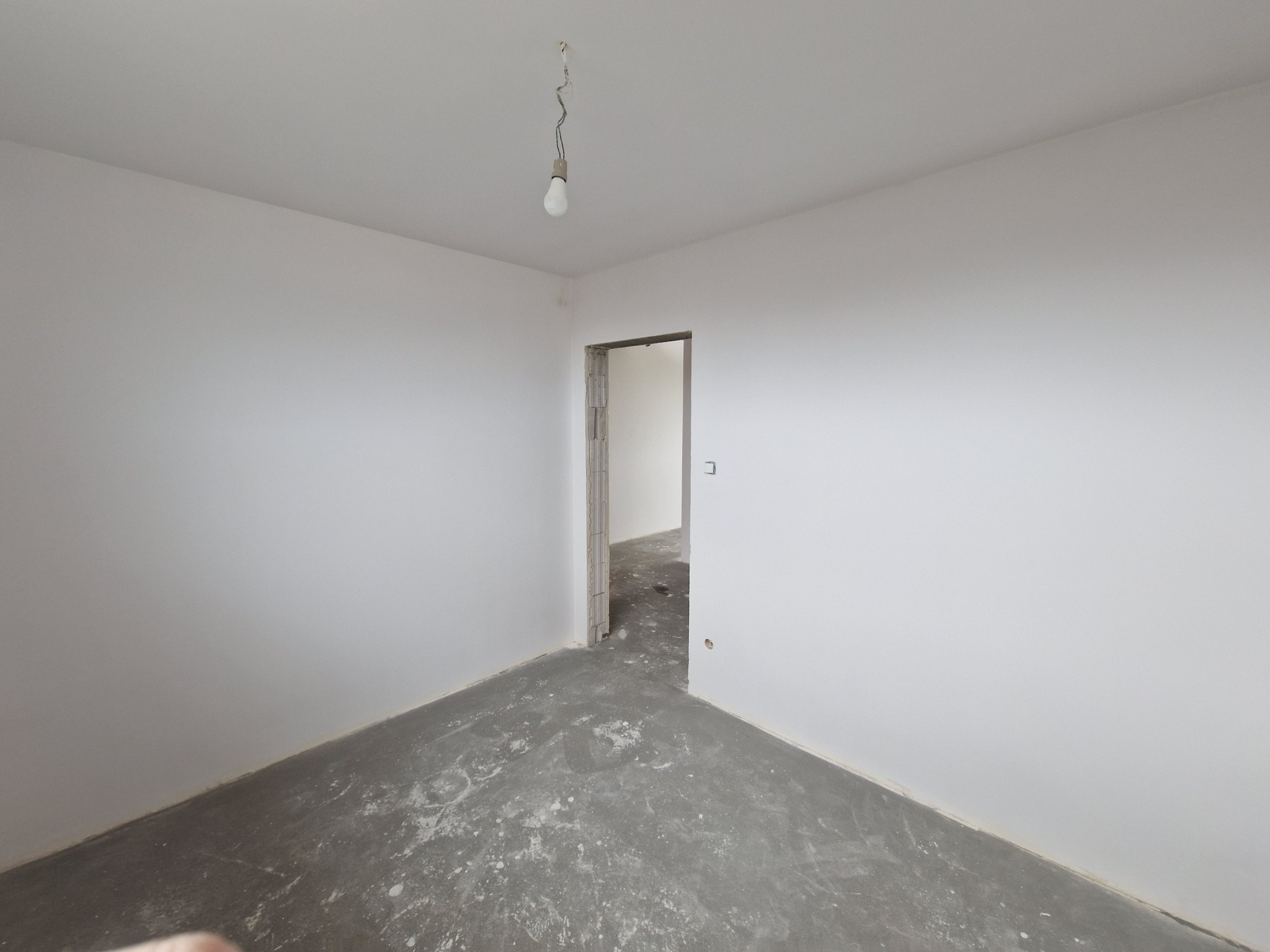Pusty pokój w trakcie remontu: białe ściany, surowy beton na podłodze, wisząca żarówka. Widoczne ślady prac wykończeniowych i przygotowania do dalszych etapów.