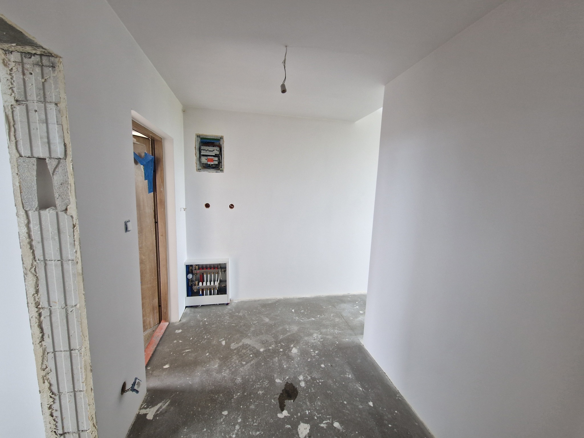 Wnętrze po szpachlowaniu i malowaniu na biało. Widoczna instalacja grzewcza, rozdzielnia elektryczna i surowa ościeżnica drzwiowa.