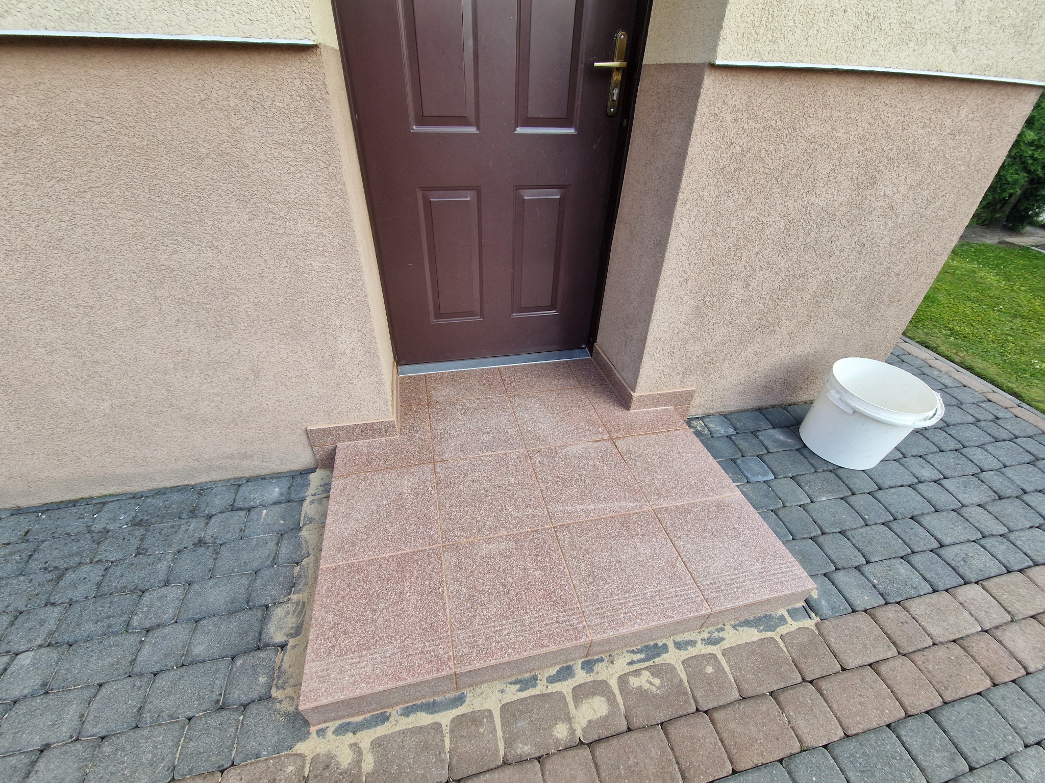 Wykończone płytkami schody wejściowe do domu, z widocznym brązowym wejściem i kostką brukową. Białe wiadro stoi obok schodów.