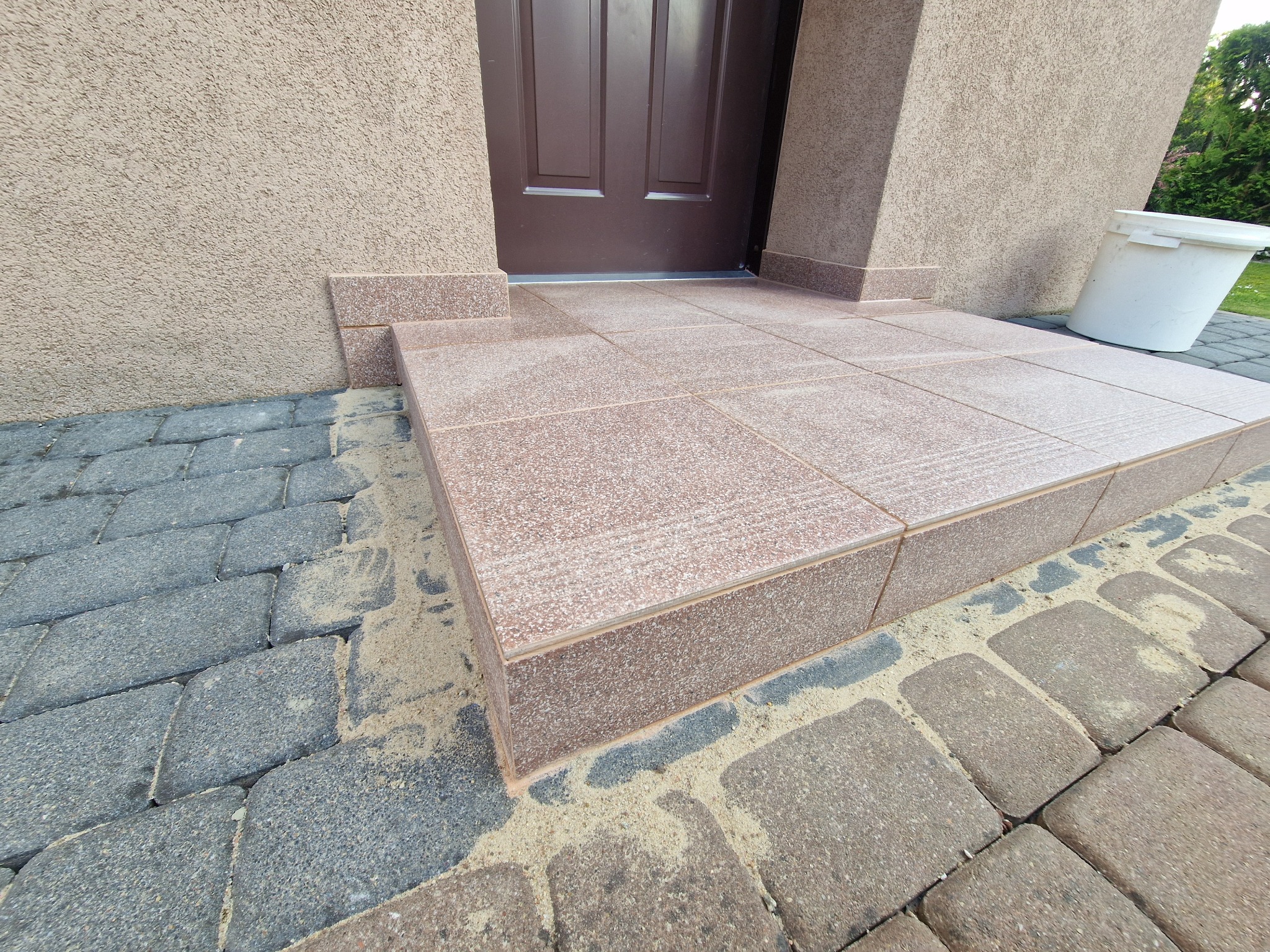 Wykończone płytkami schody wejściowe do domu, z brązowymi drzwiami w tle. Widoczne kostki brukowe wokół schodów i fragment białego wiadra.