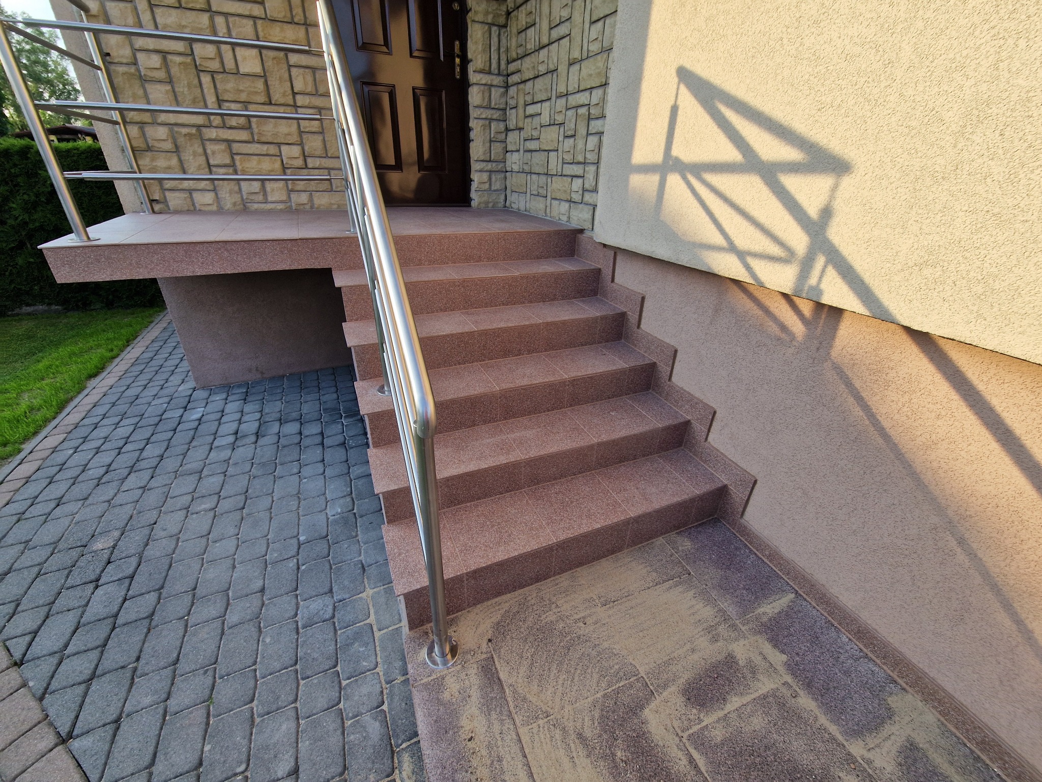 Zewnętrzne schody wejściowe wyłożone brązowymi płytkami, z metalową barierką, prowadzące do ciemnych drzwi wejściowych. Widoczna elewacja z kamienia i faktura tynku.