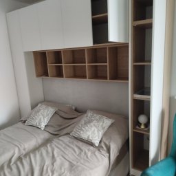 Piotr Kupryk - Biało-drewniana zabudowa sypialni z szafkami nad łóżkiem i regałem. Jasne wnętrze, beżowa pościel z wzorzystymi poduszkami. Minimalistyczny design.