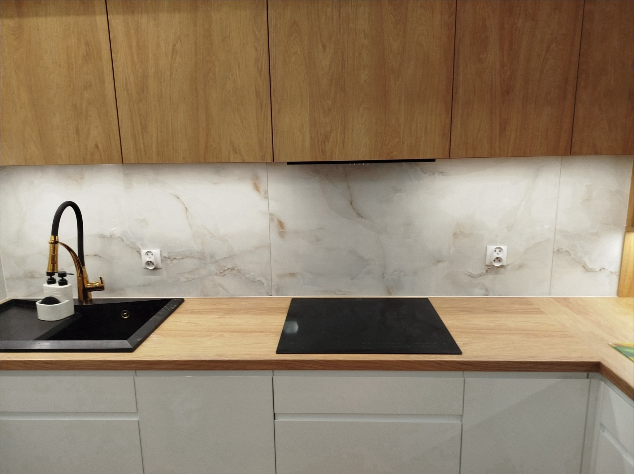 Nowoczesna kuchnia z drewnianymi szafkami górnymi i białymi dolnymi. Czarny zlew z złotą baterią, marmurowe płytki na ścianie i płyta indukcyjna.