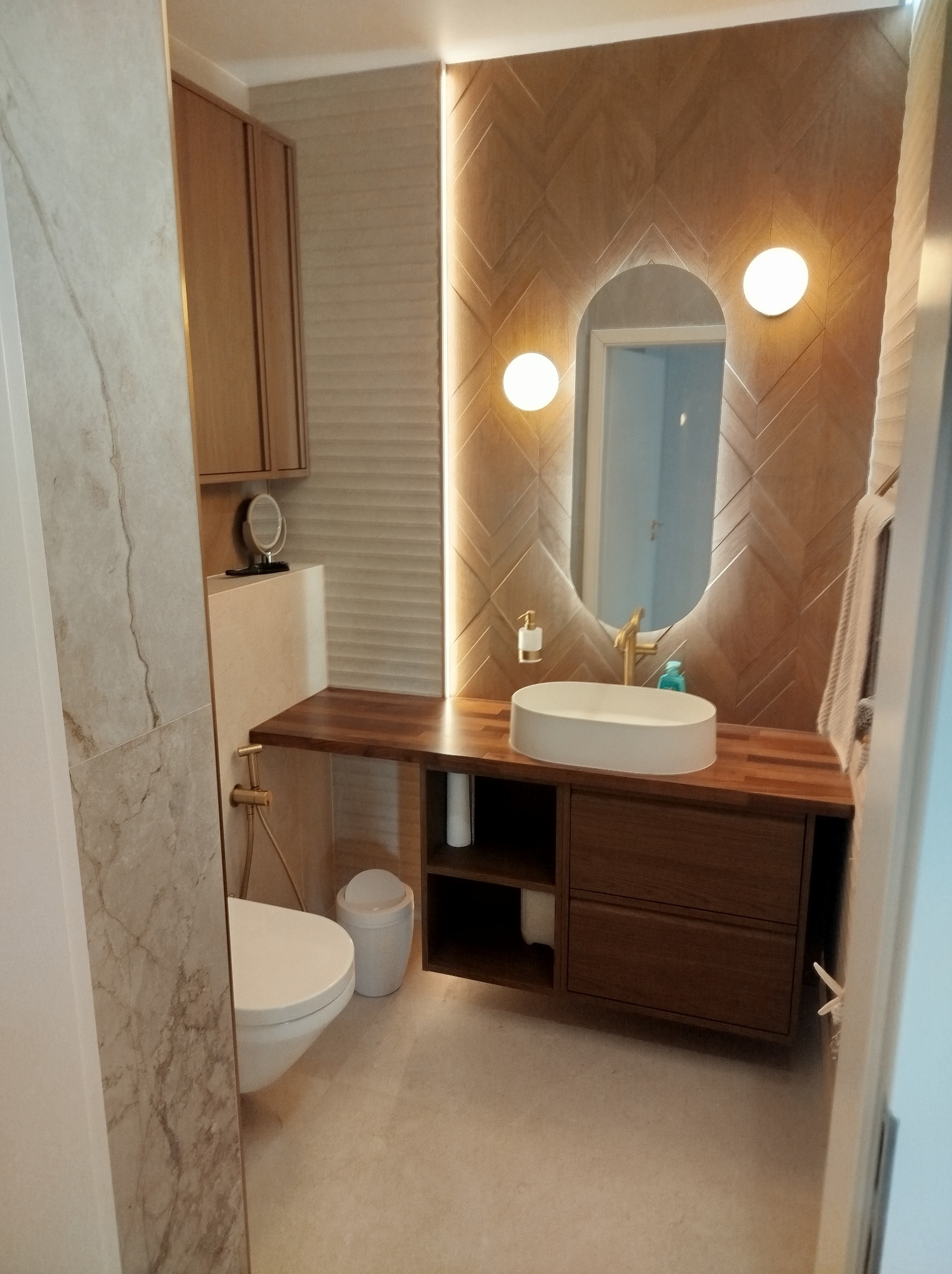 Nowoczesna łazienka z drewnianymi akcentami: szafka pod umywalką z blatem, owalne lustro z podświetleniem, toaleta i płytki w odcieniach beżu. Minimalistyczny design.