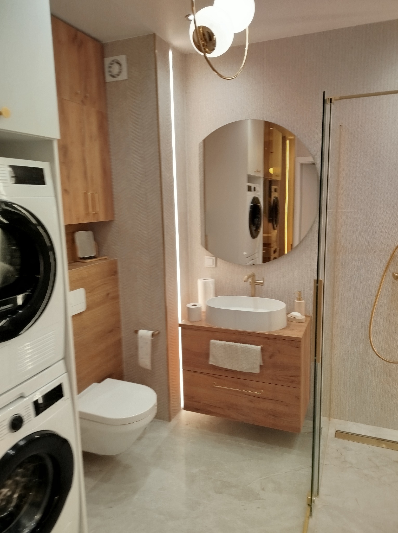 Nowoczesna łazienka z drewnianymi akcentami: szafka pod umywalką, wisząca toaleta i pralka w zabudowie. Lustro owalne odbija wnętrze. Oświetlenie punktowe.