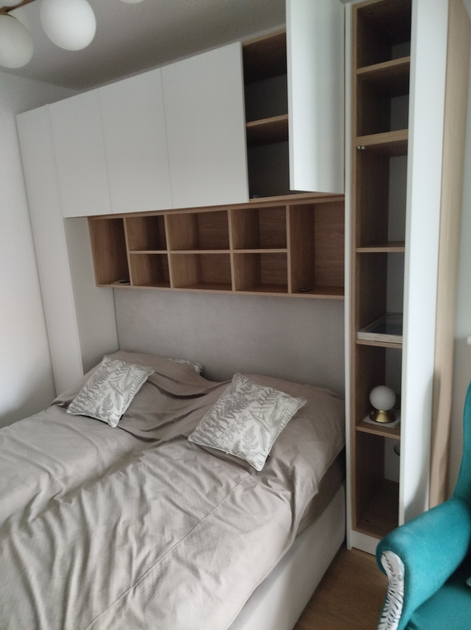 Biało-drewniana zabudowa sypialni z szafkami nad łóżkiem i regałem. Jasne wnętrze, beżowa pościel z wzorzystymi poduszkami. Minimalistyczny design.