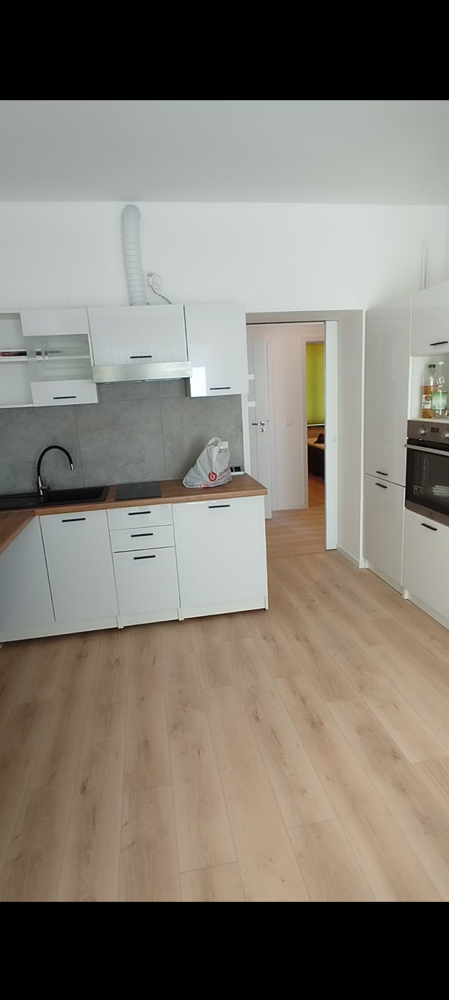 Jasna kuchnia z białymi szafkami i drewnianą podłogą. Widoczny zlew, płyta grzewcza i okap. W tle otwarte drzwi do innego pomieszczenia.