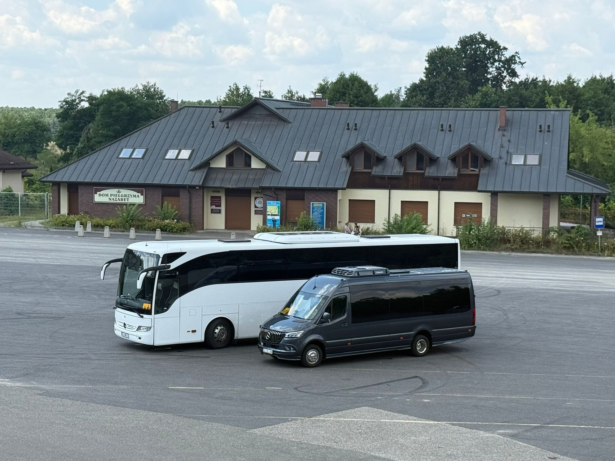 Autokary i bus w Głownie, zaparkowane przed budynkiem Domu Pielgrzyma. Biały autokar i ciemny bus z przyciemnianymi szybami.