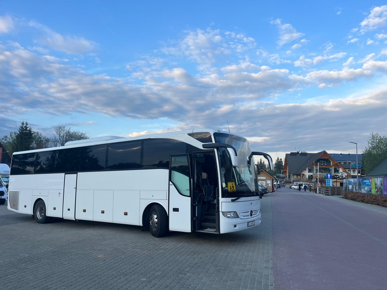Nowoczesny, biały autokar Mercedes z otwartymi drzwiami na tle górskiego krajobrazu i błękitnego nieba z chmurami. Widoczny znak z sylwetkami dzieci.