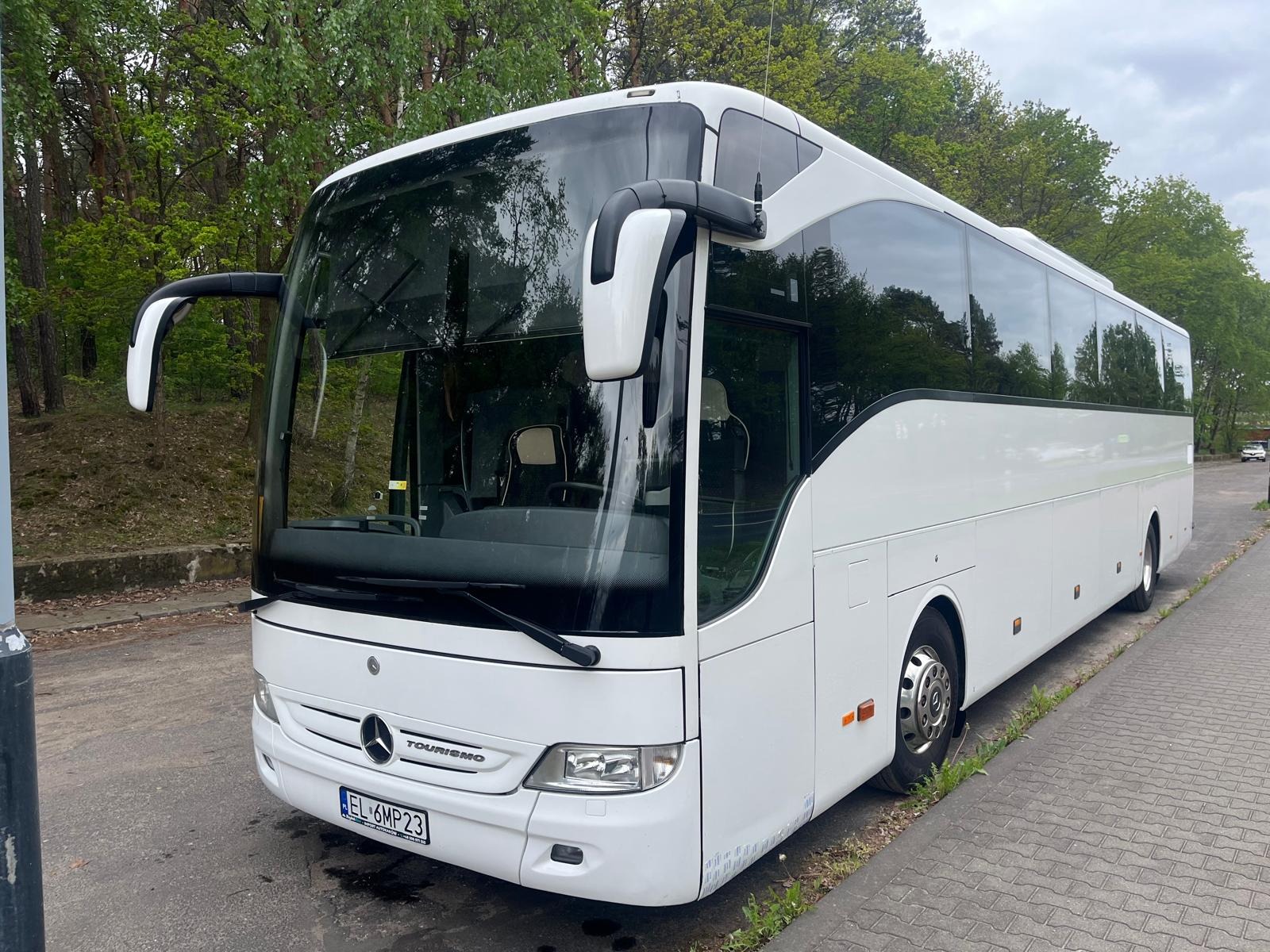 Nowoczesny, biały autokar Mercedes-Benz Tourismo zaparkowany na tle zieleni. Widoczny z przodu, z numerem rejestracyjnym EL 6MP23. Autobus czysty, zadbany, gotowy do trasy.