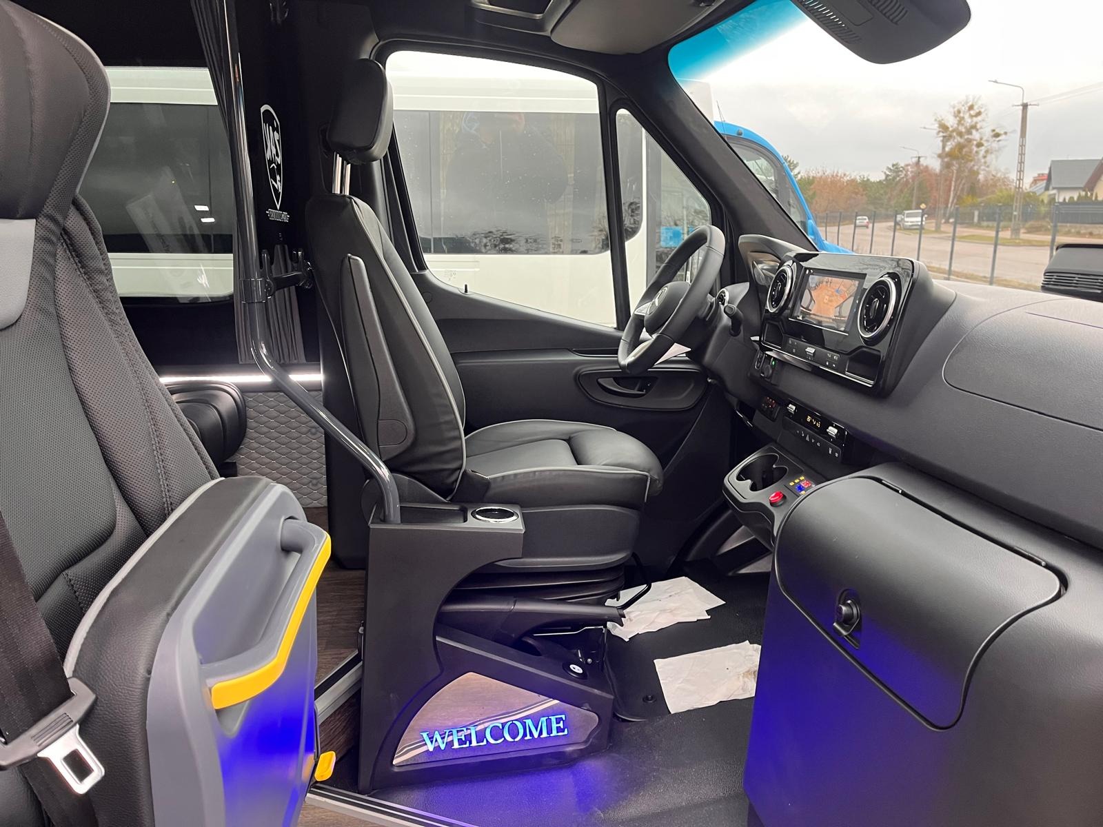 Wnętrze luksusowego busa z skórzanymi fotelami, oświetleniem LED i napisem 'WELCOME'. Widok na kokpit, kierownicę i system multimedialny. Detale wykończenia premium.