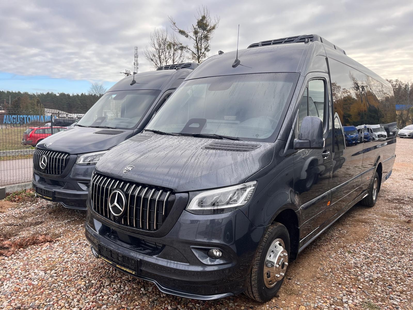 Dwa ciemne busy Mercedes-Benz Sprinter z chromowanymi grillami, zaparkowane na żwirowym parkingu w pochmurny dzień. Widoczny napis 'Augustów' na ogrodzeniu w tle.