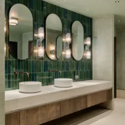 Łazienka z trzema owalnymi lustrami nad umywalkami, zielone płytki na ścianie i drewniane szafki pod blatem z białego marmuru, nowoczesne oświetlenie.