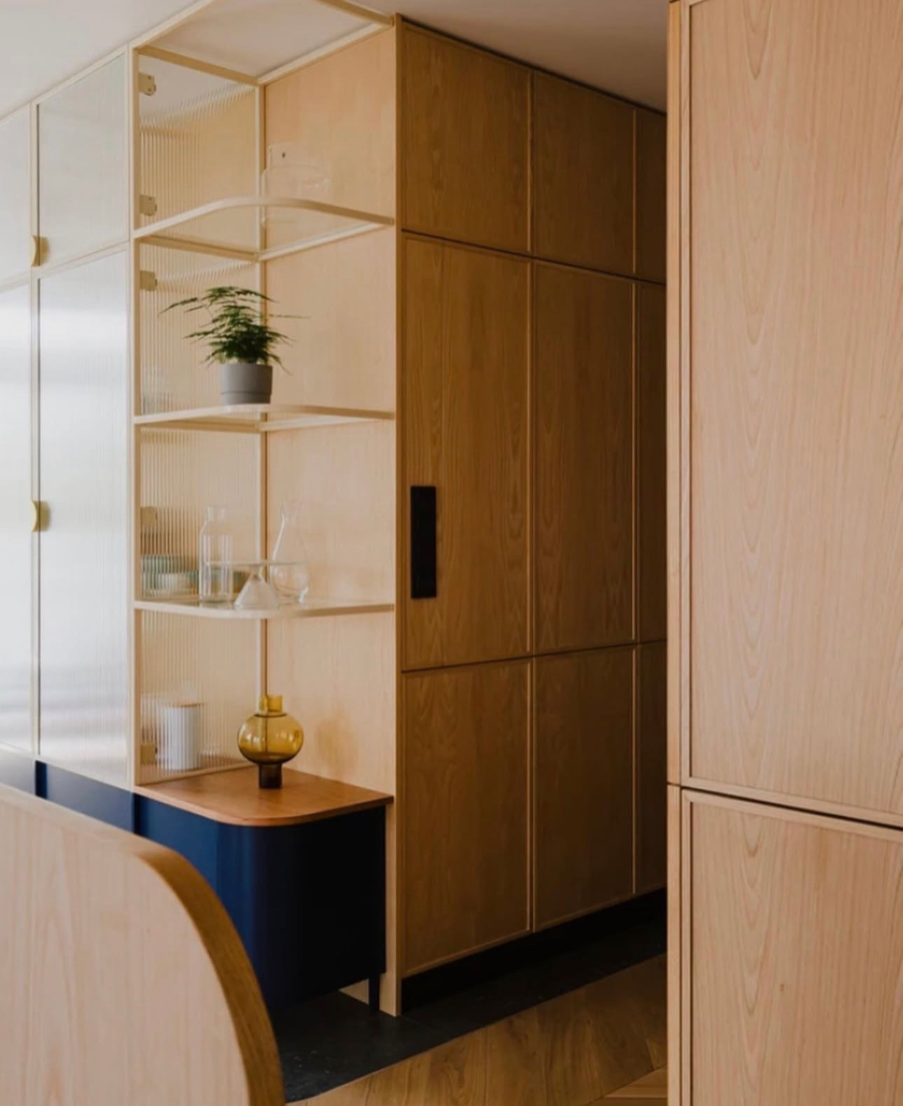 Drewniana zabudowa wnęki z półkami z ryflowanego szkła, uzupełniona o szafki z ciemnymi uchwytami. Na półkach dekoracje i roślina doniczkowa. Ujęcie z bliska.