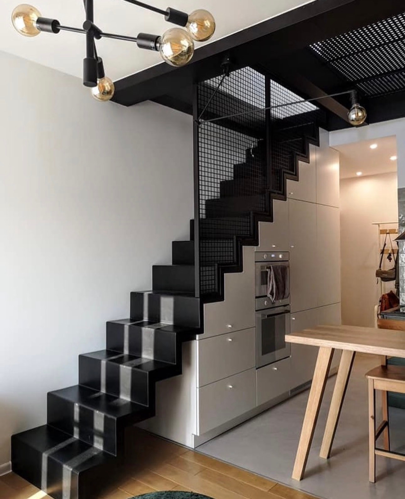 Nowoczesna kuchnia z zabudową pod schodami, czarne schody z szarymi akcentami, oświetlenie punktowe i drewniany stół. Minimalistyczny design.
