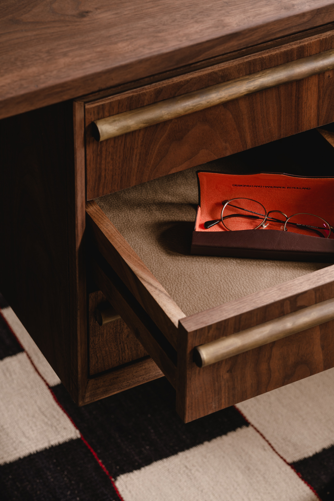 Otwarta szuflada biurka z orzecha amerykańskiego z mosiężnymi uchwytami, wewnątrz etui na okulary. Na podłodze wzorzysty dywan.