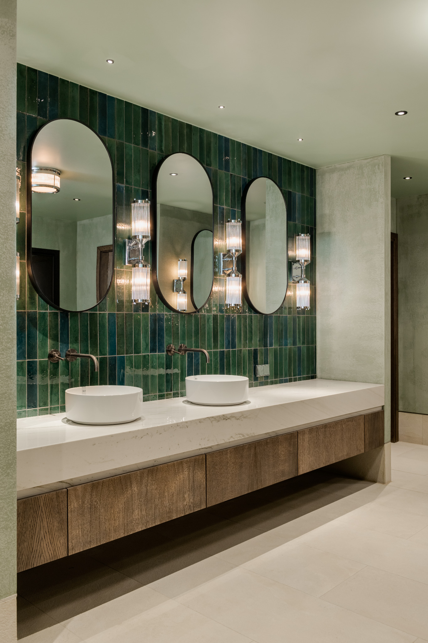 Łazienka z trzema owalnymi lustrami nad umywalkami, zielone płytki na ścianie i drewniane szafki pod blatem z białego marmuru, nowoczesne oświetlenie.