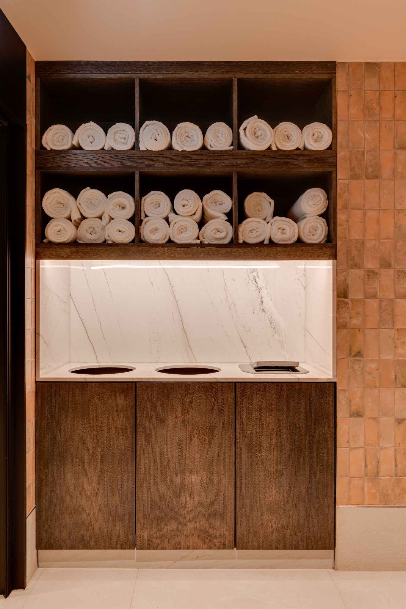 Drewniana szafka z marmurowym blatem i wnękami na ręczniki w łazience. Oświetlenie LED podkreśla teksturę marmuru i ciepłą kolorystykę płytek ściennych.