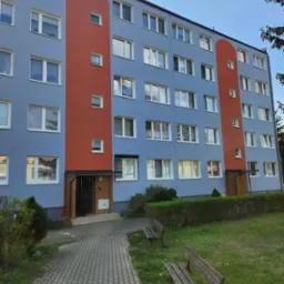 Odnowiona fasada bloku w błękitno-pomarańczowych barwach, z widoczną ścieżką i ławkami. Estetyczne wykończenie budynku.