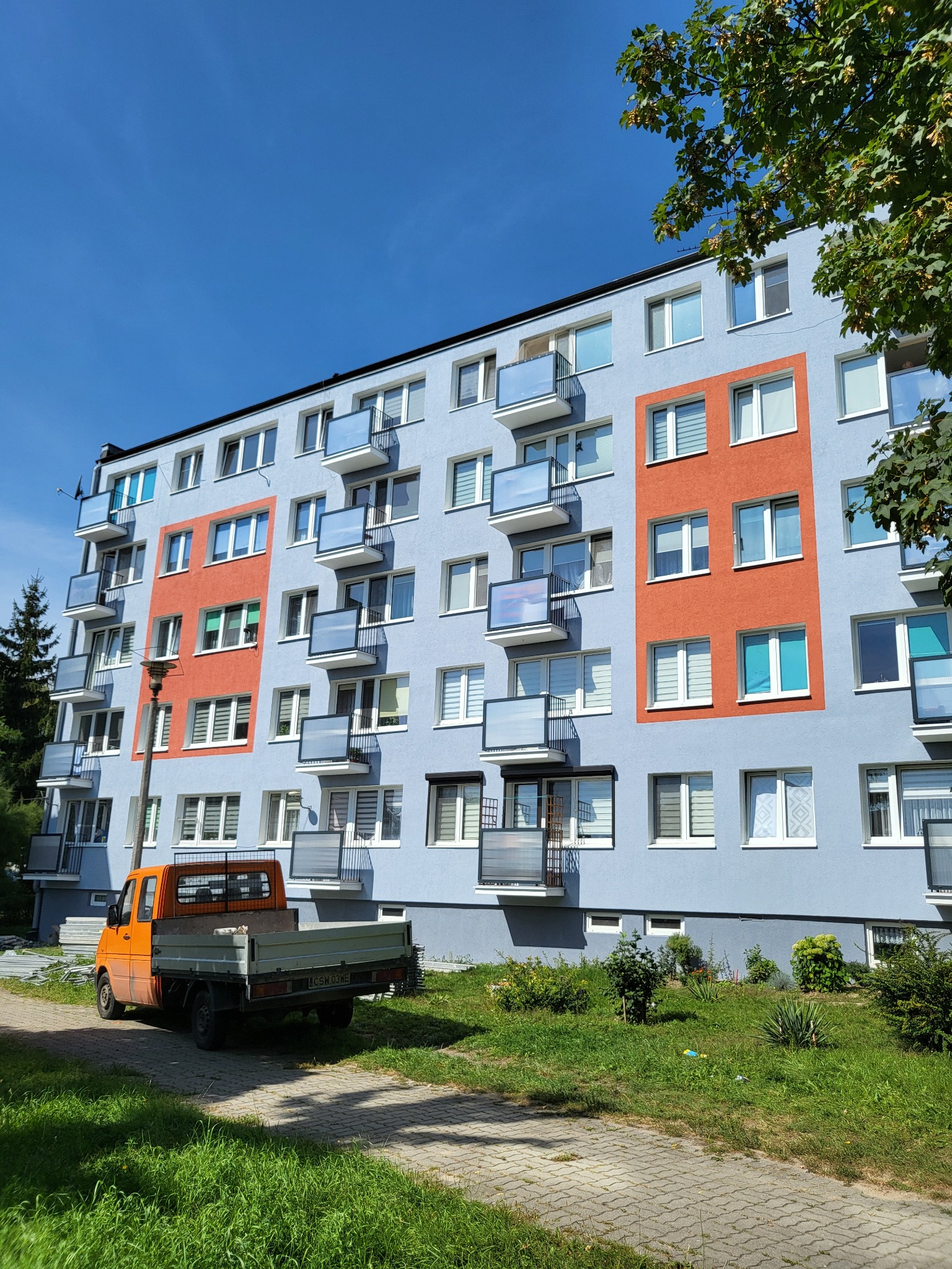 Wielopiętrowy budynek po renowacji elewacji, z pomalowanymi na pomarańczowo fragmentami. Na pierwszym planie pomarańczowy samochód dostawczy.