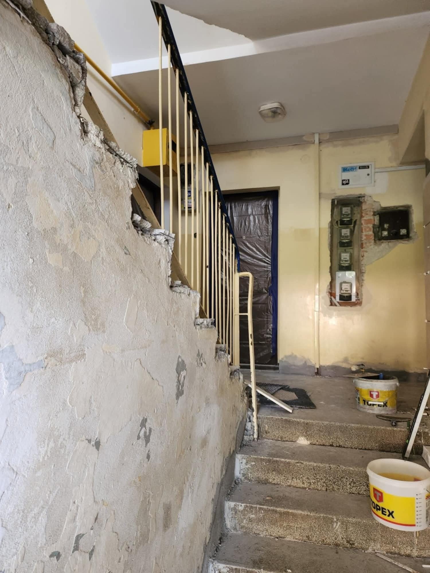 Klatka schodowa w trakcie remontu: uszkodzona ściana obok schodów, odsłonięte przewody elektryczne i liczniki, widoczne wiadra z farbą, przygotowane do prac wykończeniowych.