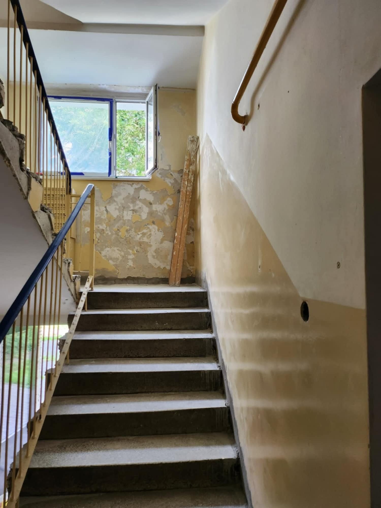 Klatka schodowa w trakcie prac. Odkryte warstwy farby na ścianie, widoczne schody i okno z zielenią na zewnątrz. Fragment poręczy i uszkodzone fragmenty ścian.