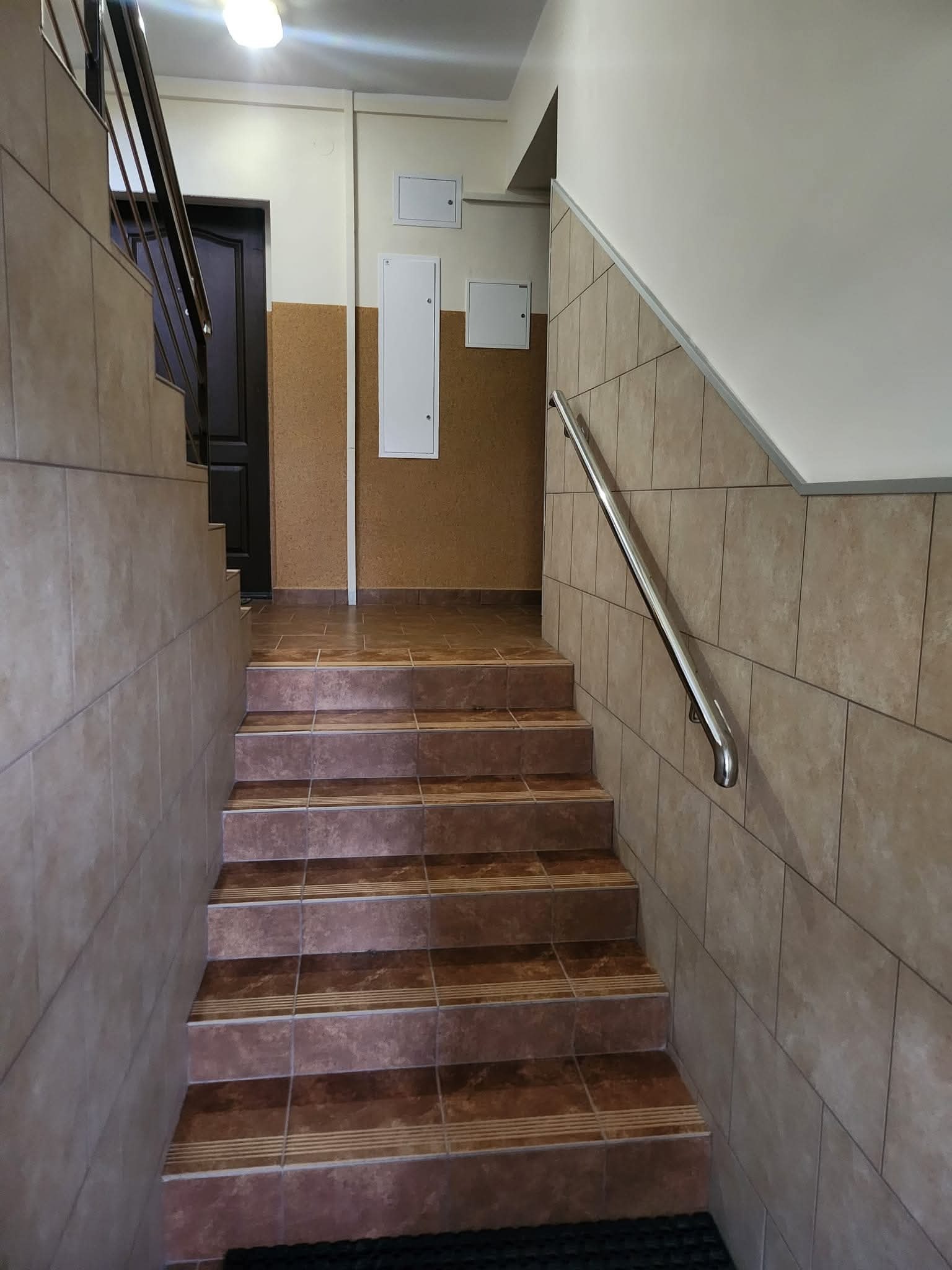 Klatka schodowa z brązowymi płytkami, wykończona metalową poręczą. Widoczne schody, ściany wyłożone płytkami i drzwi w tle. Oświetlenie sufitowe.