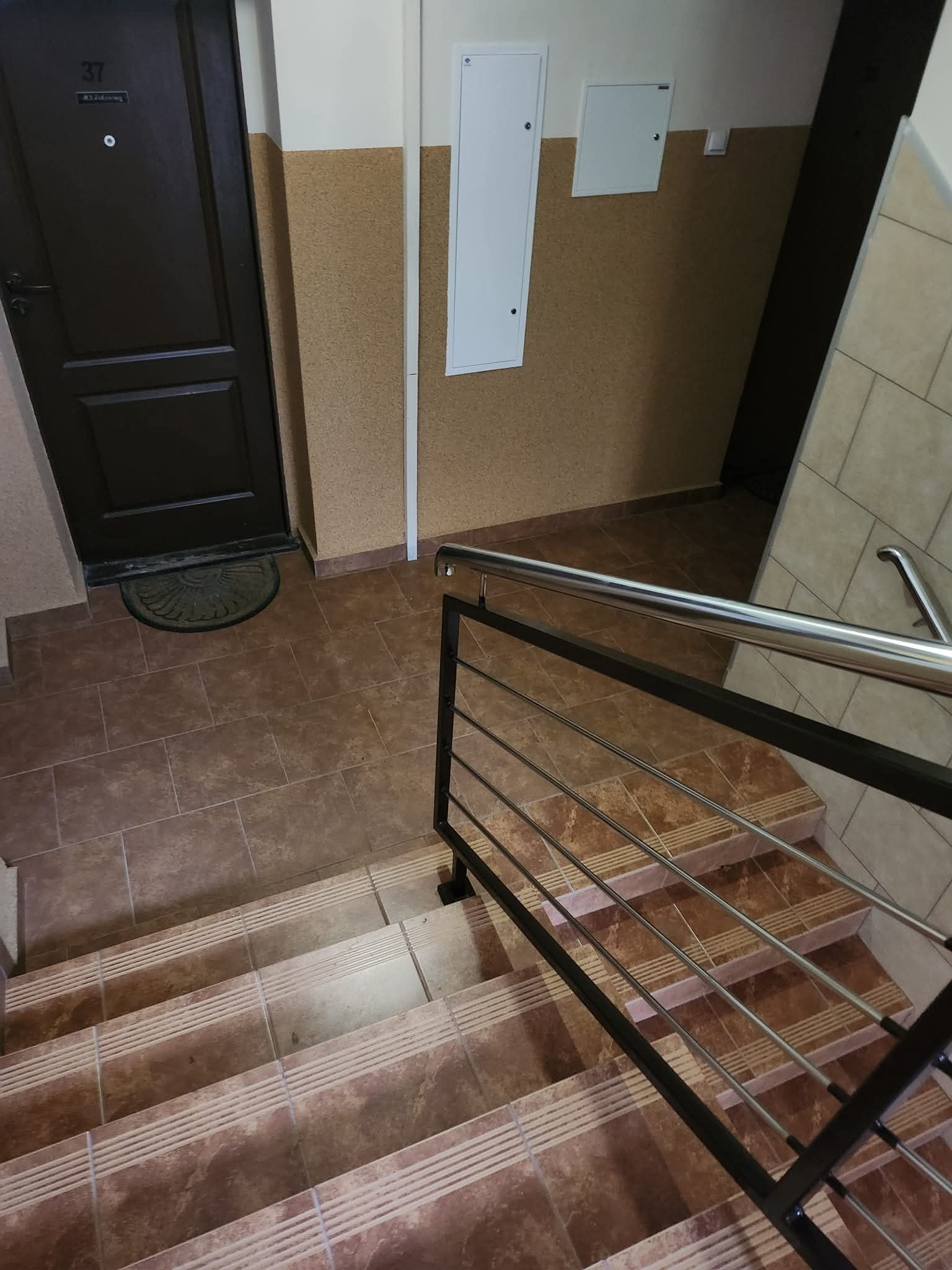 Klatka schodowa w budynku wielorodzinnym z brązowymi płytkami na schodach i podłodze. Widoczne drzwi do mieszkania z numerem 37 i metalowa poręcz.