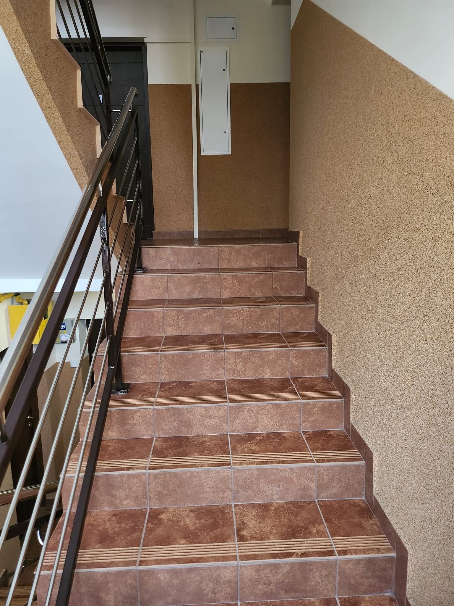 Wykończone schody w budynku wielorodzinnym, brązowe płytki z paskami antypoślizgowymi, pomalowane ściany z fakturą, metalowa barierka. Klatka schodowa po remoncie.