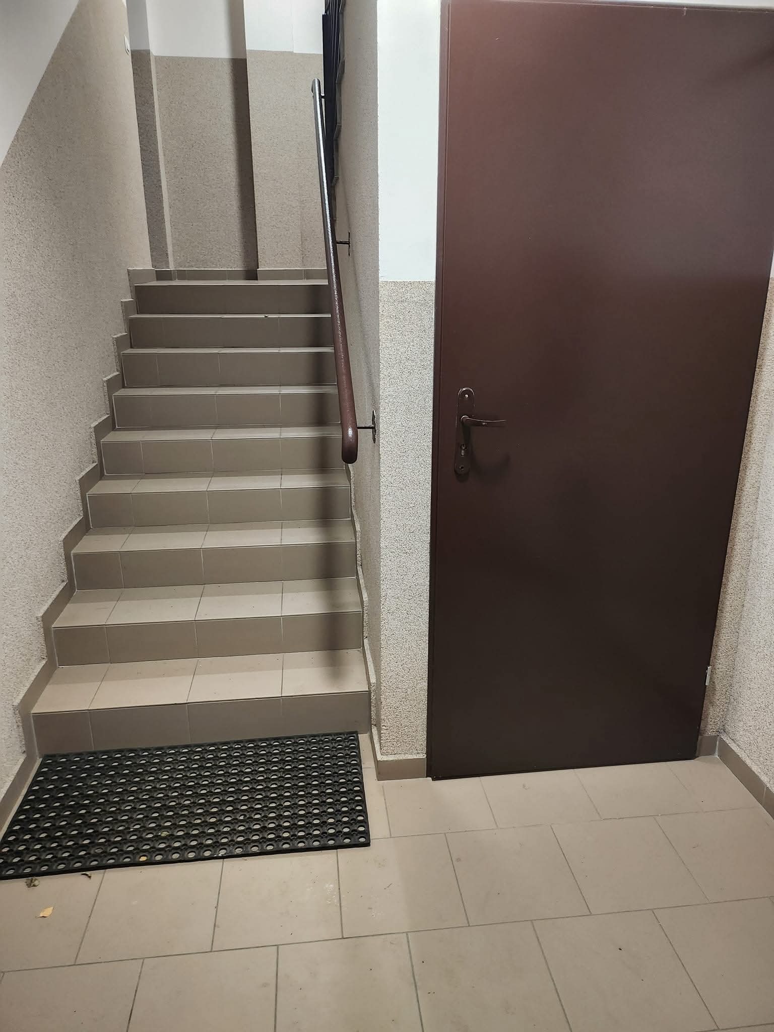 Klatka schodowa z brązowymi drzwiami i szarymi schodami wyłożonymi płytkami. Na podłodze wycieraczka. Ściany pokryte tynkiem strukturalnym.