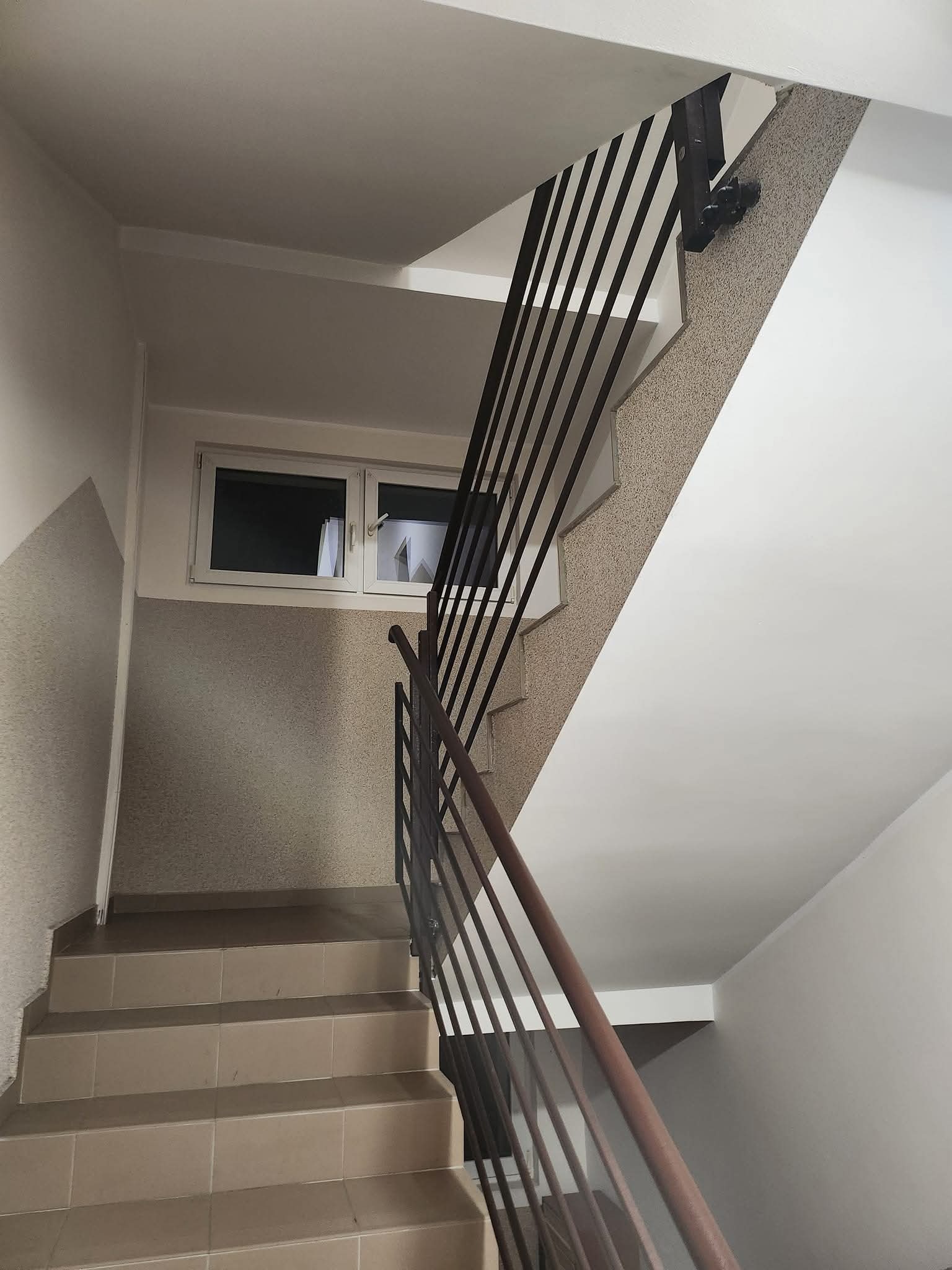 Klatka schodowa po remoncie: jasne ściany z fakturą, brązowe płytki na schodach i metalowa balustrada. Widoczne okno na półpiętrze, oświetlenie sufitowe.