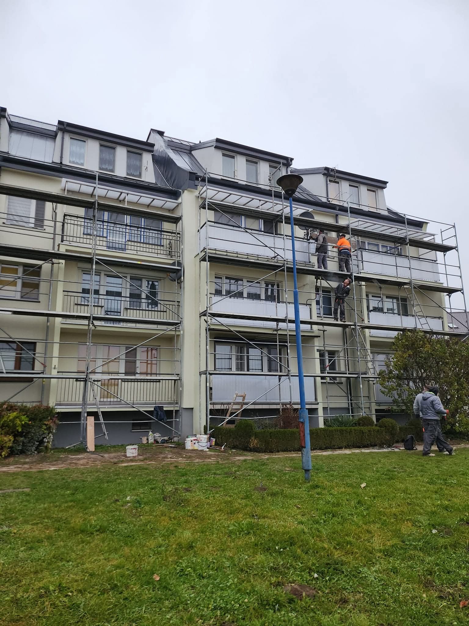 Elewacja budynku w trakcie malowania z rusztowania. Trzech malarzy przy pracy, farby i narzędzia na trawniku, widoczny fragment lampy ulicznej.