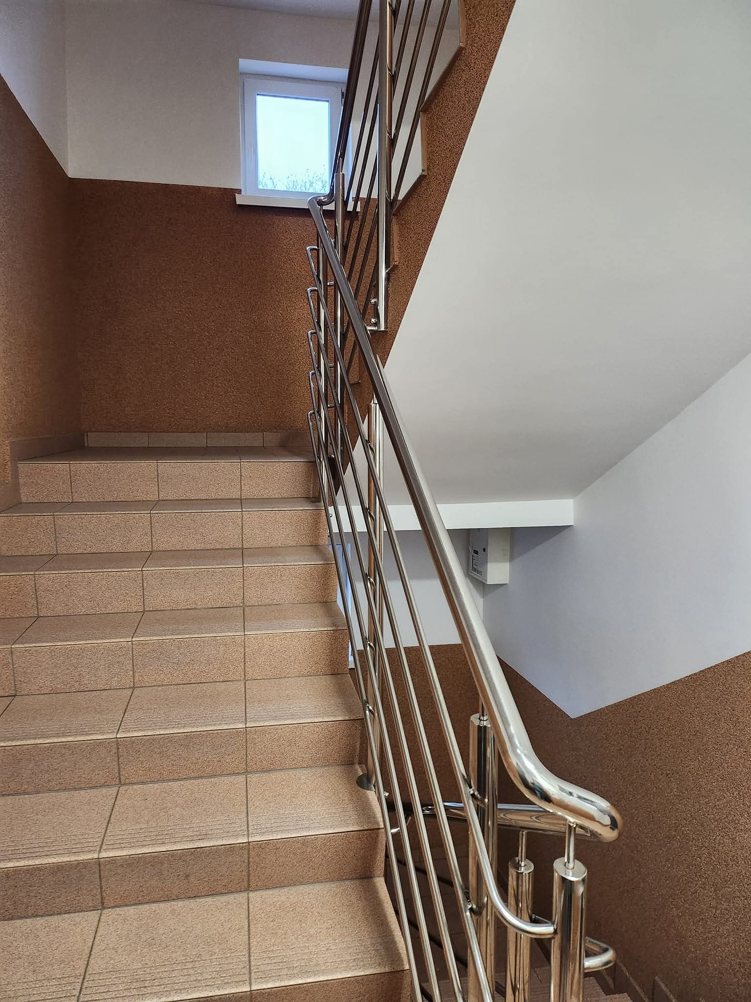 Klatka schodowa wykończona brązowymi płytkami i stalową balustradą. Widoczne okno na półpiętrze i fragment białego sufitu. Teksturowana ściana w kolorze brązu.