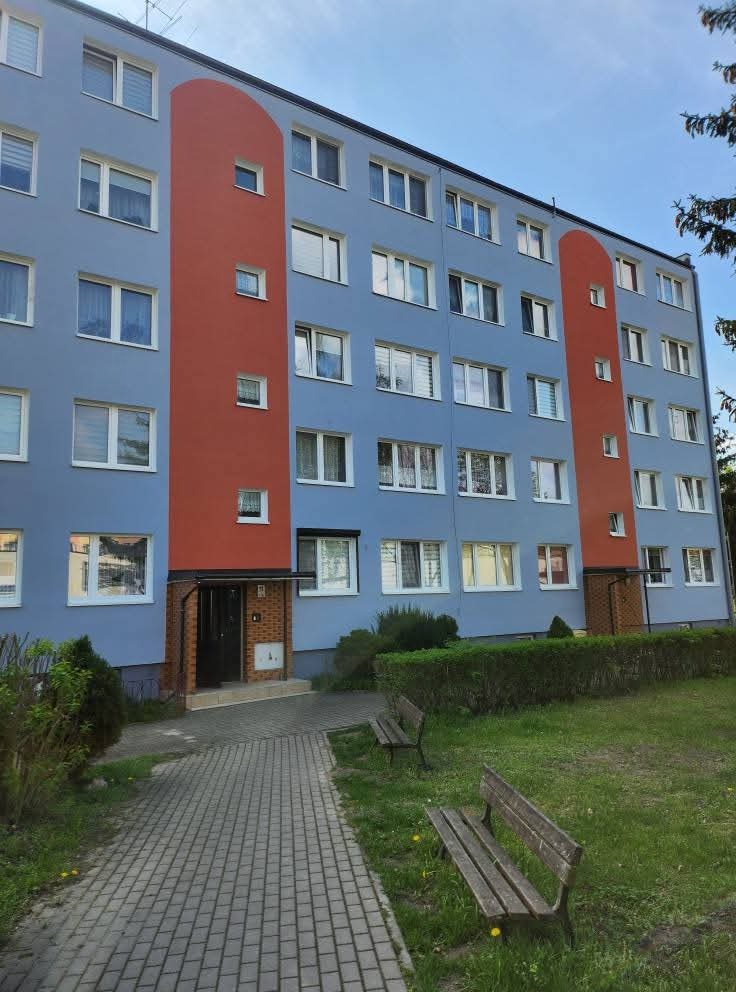 Odnowiona fasada bloku w błękitno-pomarańczowych barwach, z widoczną ścieżką i ławkami. Estetyczne wykończenie budynku.