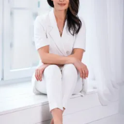 Portret kobiety w białym garniturze, siedzącej na tle okna. Uwagę przyciąga elegancki strój i stonowana kolorystyka zdjęcia. Całość utrzymana w jasnej tonacji.