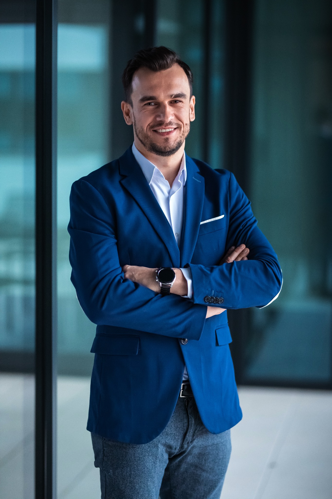 Uśmiechnięty mężczyzna w błękitnej marynarce, z zegarkiem na ręku, pozuje na tle szklanej ściany we Wrocławiu. Zdjęcie biznesowe.