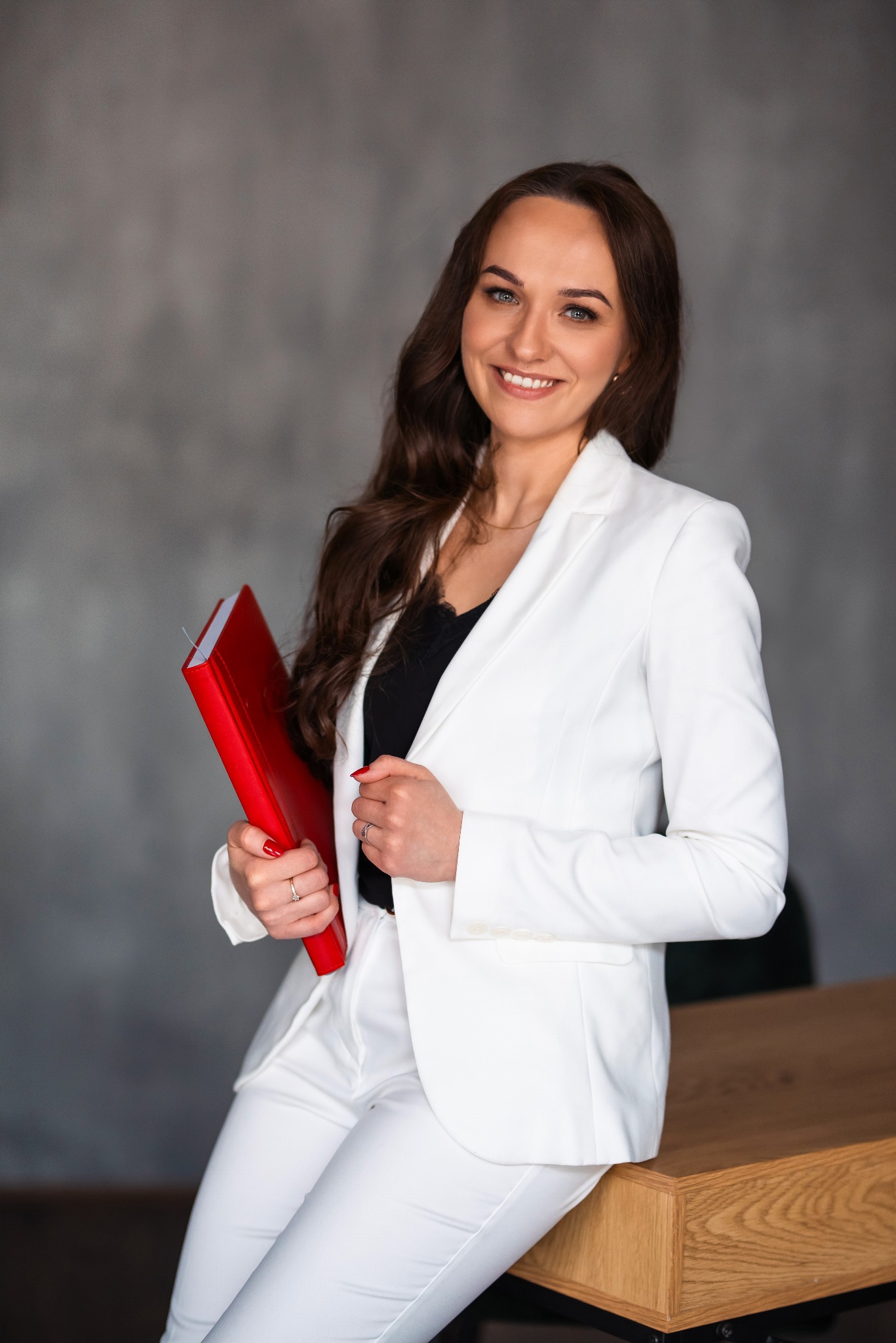 Uśmiechnięta brunetka w białym garniturze, trzymająca czerwoną teczkę, opiera się o biurko. Zdjęcie biznesowe w studio z szarym tłem.