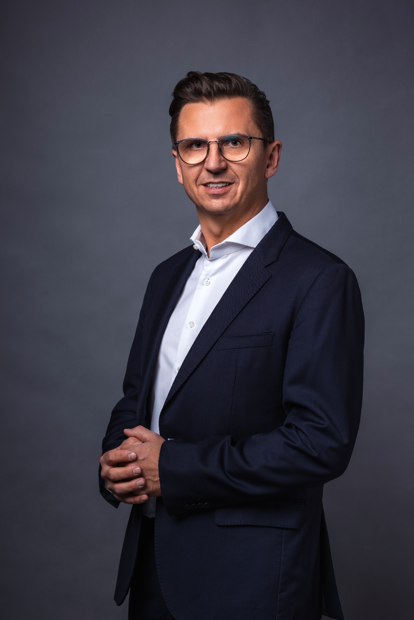 Mężczyzna w okularach, białej koszuli i granatowej marynarce, z dłońmi splecionymi przed sobą, na szarym tle. Portret biznesowy.